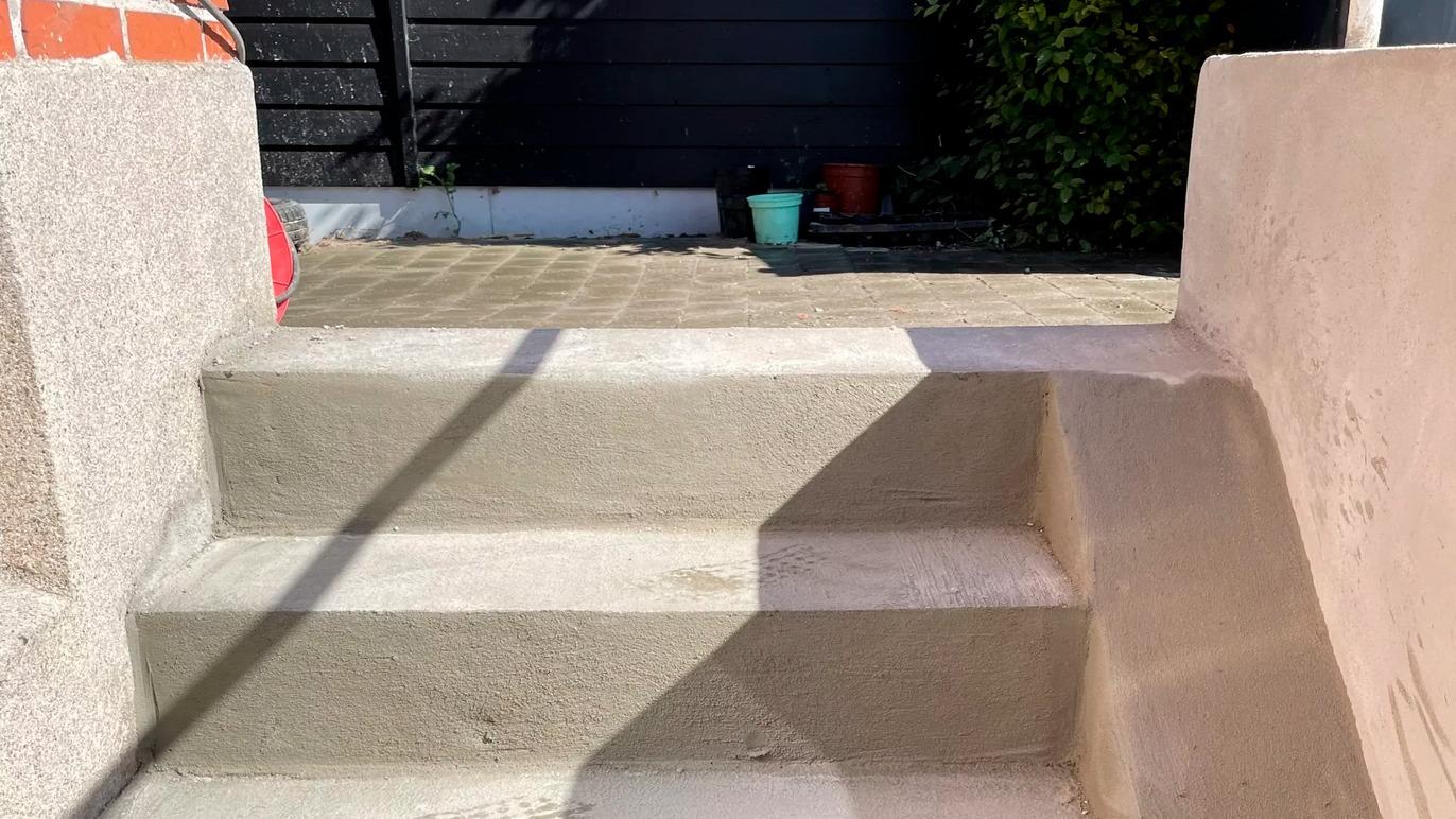 Trappe med tre trin, belagt med lys cement, fører op til en gård med grønne planter i baggrunden.