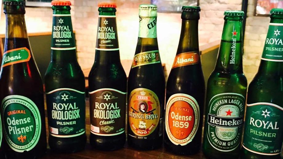 Flere forskellige ølflasker, herunder Odense Pilsner, Royal Biologisk og Heineken, præsenteret på en bar.
