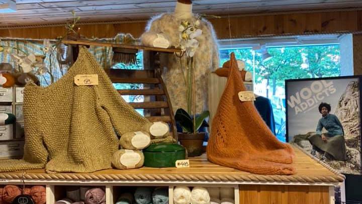 Håndstrikkede tæpper og garnnøgler præsenteret i en hyggelig butik med træhylder og vindue i baggrunden.