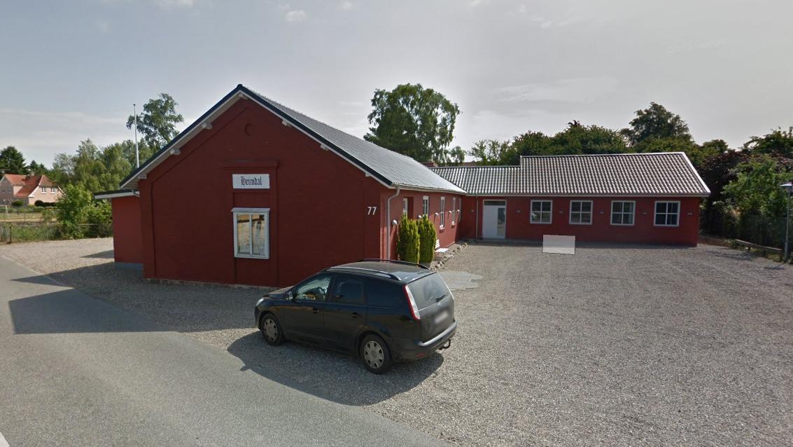 Rød bygning med grå tag og parkeret bil på grusvej i et roligt område.