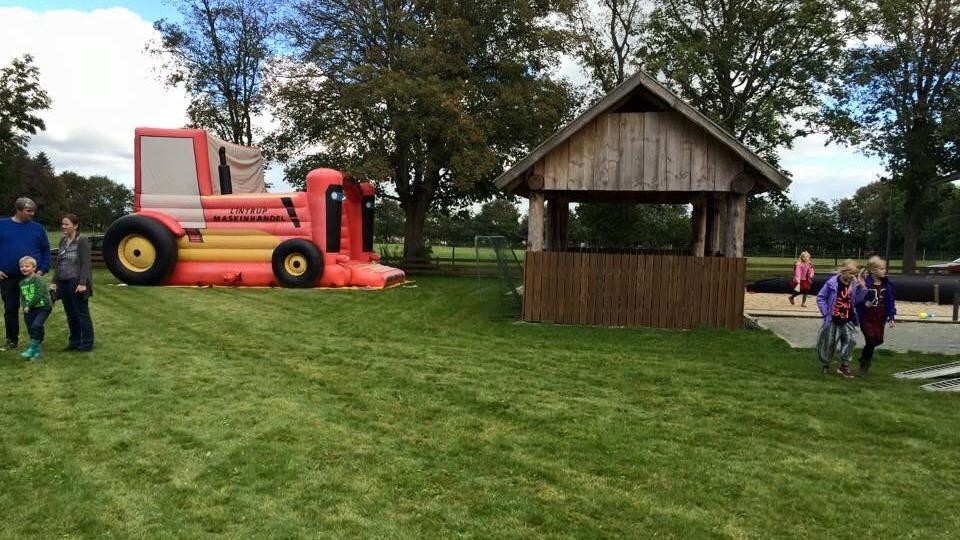 Børnevenlig legeplads med en oppustelig traktor og en træhytte omgivet af grøn græsplæne og glade børn.