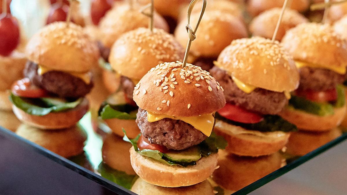 Små hamburgere med sesamfrø, salat, tomat og ost præsenteret på en spejlplade.
