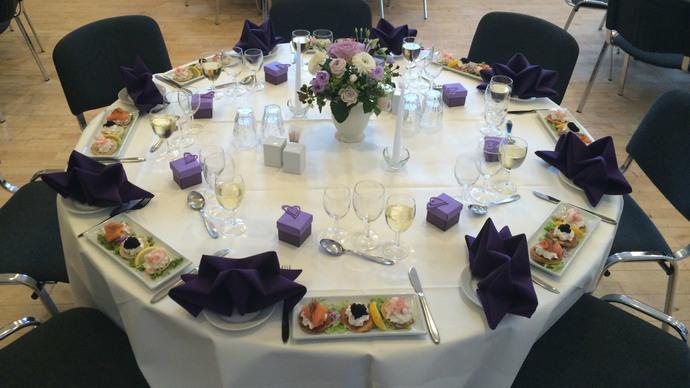 Elegant dækket bord med lilla servietter, sushi og blomsterarrangement i midten.