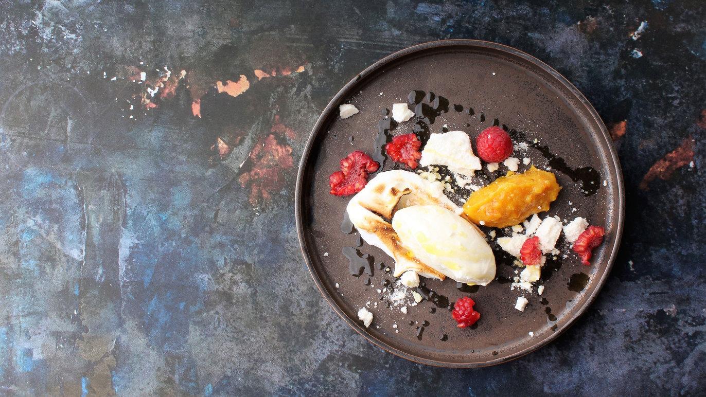 En tallerken med dessert bestående af is, hindbær, mango og smuldret hytteost på en farverig baggrund.