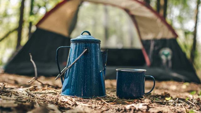 Blå kaffekande og krus på jorden foran et telt i skoven.
