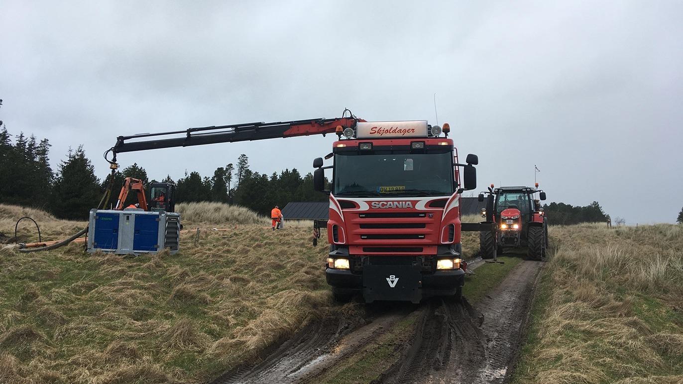 En rød lastbil med kran arbejder på en mark, mens en traktor kører på en mudret vej.