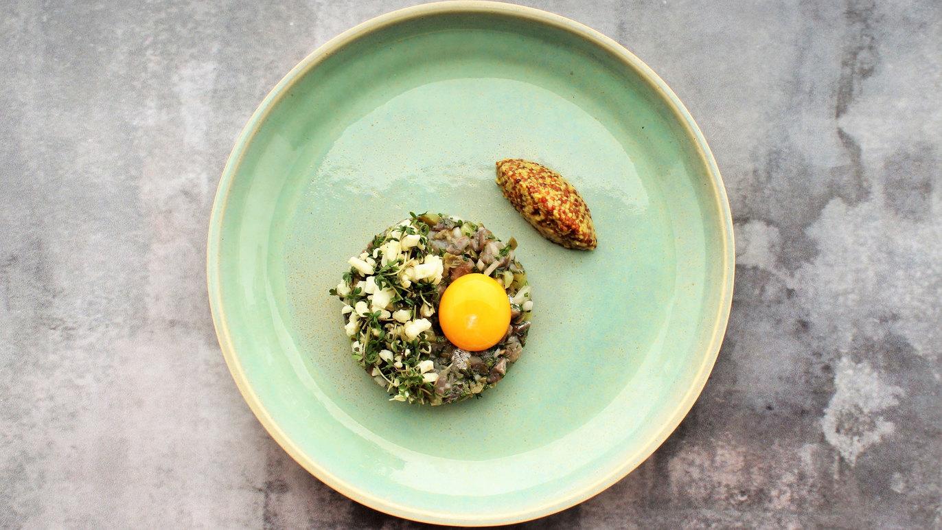 En tallerken med en portion tartar, et æggeblomme og en sprød agurk på en lysgrå overflade.