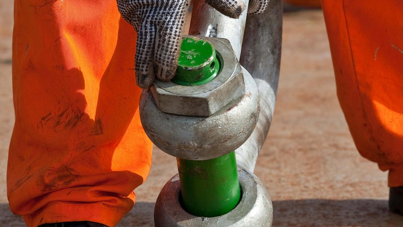 Hånd, der justerer en grøn metalforbindelse med en søm og beskyttelseshandsker, iført orange arbejdstøj.