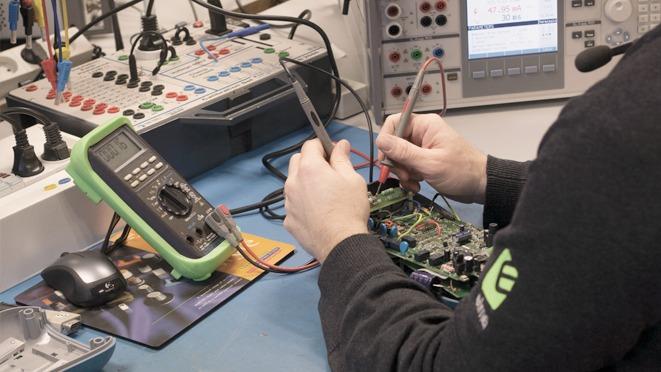 En tekniker arbejder med en multimeter og måler på et kredsløb på et værksted med elektronisk udstyr.