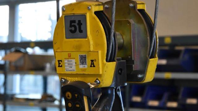 En gul kran med en kapacitet på 5 ton, placeret i et lagerområde med hylder i baggrunden.