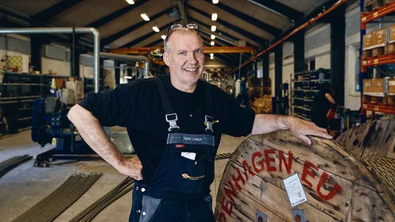 En smilende mand i arbejdsbukser står ved siden af en stor træplade i en travl værksted.