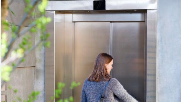 En kvinde står foran en elevator med metaldøre og trykker på knappen for at åbne den.