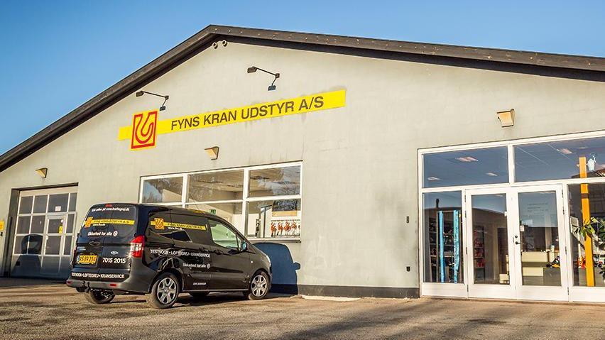 Bygning med skilt for Fyns Kran Udstyr A/S og en varevogn parkeret foran indgangen.