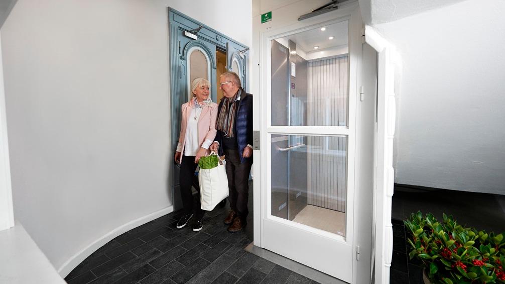 To personer, en kvinde med en taske og en mand, står ved en elevator i en moderne bygning.