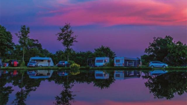 Campingvogne og biler ved en sø med farverige skyer i solnedgangen, der spejler sig i vandet.