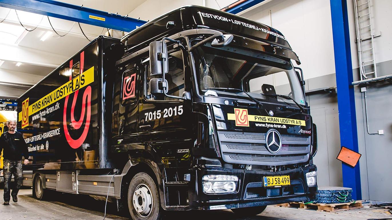 En sort Mercedes lastbil med logoer og tekst, parkeret i en garage med blå vægge og en løftekran.