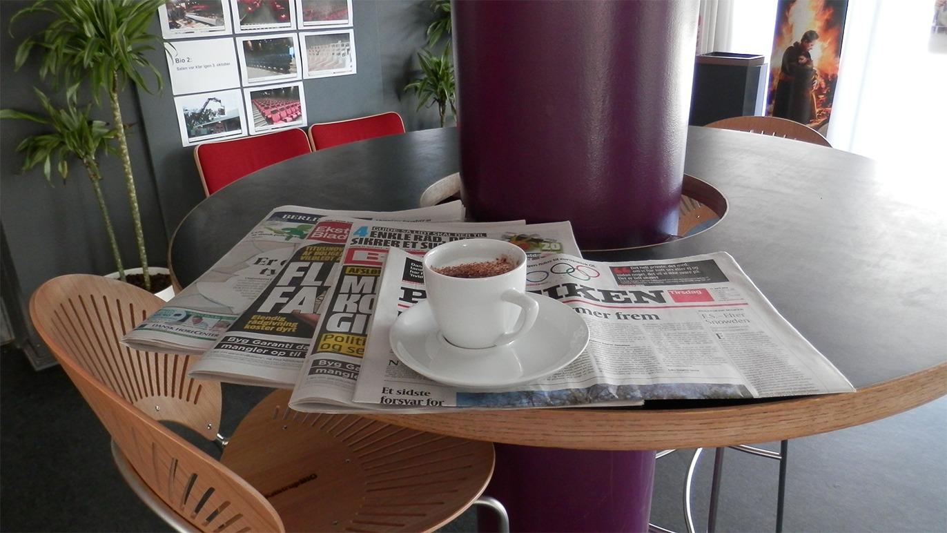 En kop kaffe på en hvid tallerken, der står på et bord med aviser i en café.