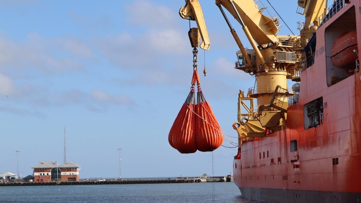 Et skib løfter en orange redningskrans fra havnen under en klar himmel.