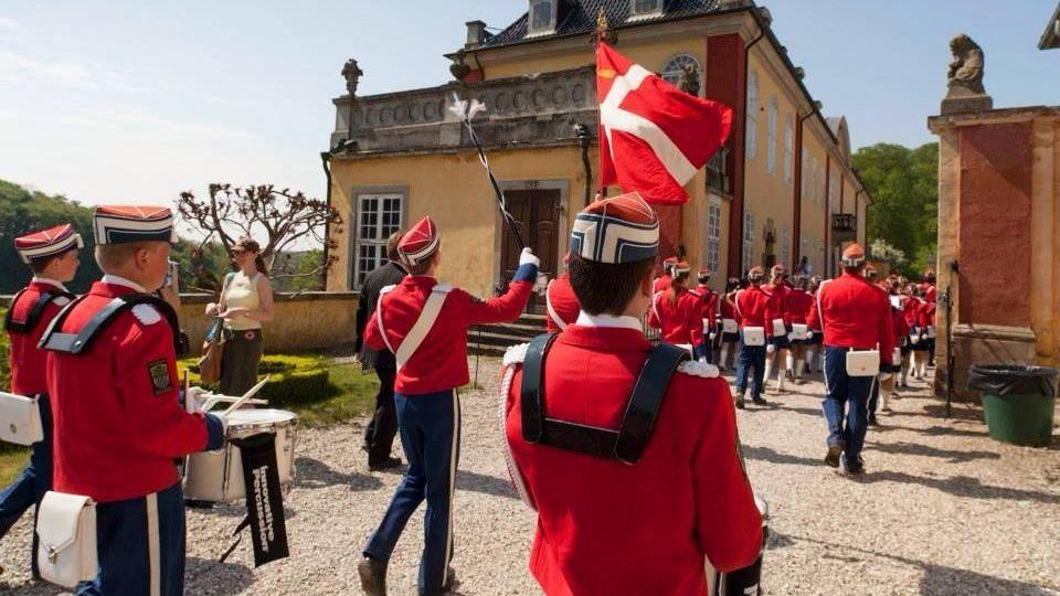 En parade med musikere i røde uniformer, der bærer det danske flag foran en historisk bygning.