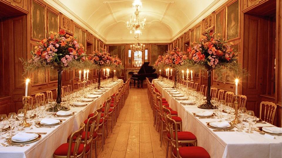 Elegant spisestue med lange borde dækket til fest, blomsterdekorationer og stearinlys i en historisk atmosfære.