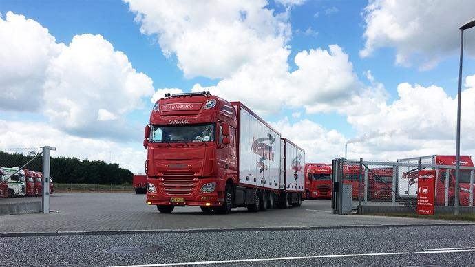 Rød lastbil med to trailere parkeret på en rasteplads under en blå himmel med skyer.