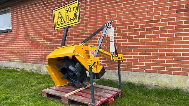 Gul maskine til jordbearbejdning placeret på en palle foran en murstensvæg med advarselsskilt.