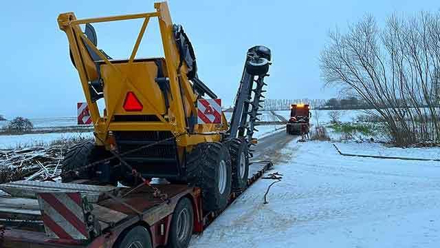 En gult maskine med store dæk står på en trailer i sneen, mens en anden maskine kører på vejen.