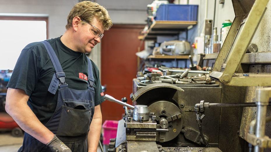 En mekaniker arbejder koncentreret ved en drejebænk i et værksted, omgivet af værktøj og maskiner.