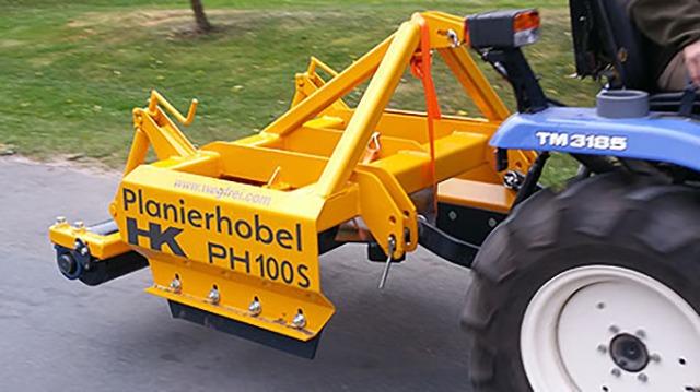 En gul planihobel monteret på en traktor, der kører på en grusvej.