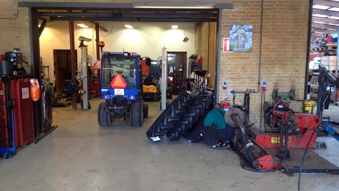 Værksted med maskiner, dæk og mekanikere, der arbejder på en bil i en garage med værktøj og udstyr.