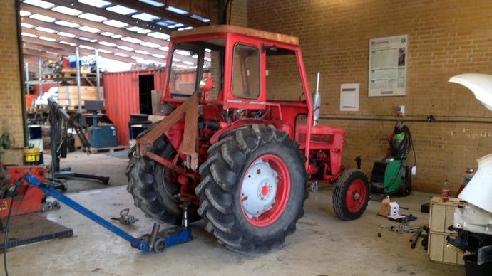 Rød traktor i en værksted med værktøj og udstyr omkring, klar til reparation.
