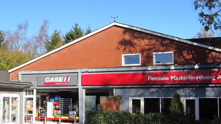 Facade af en butik med skiltet "CASE" og "Remlase Maskinforretning" under en klar blå himmel.
