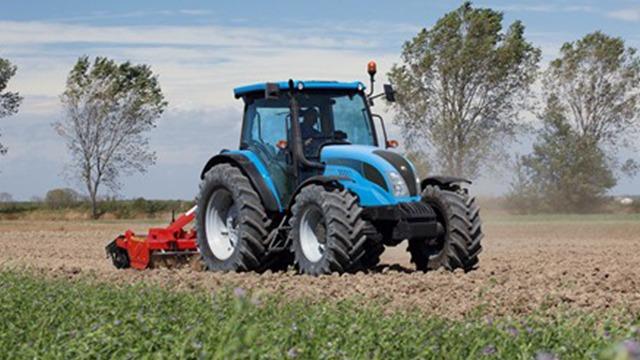 En blå traktor arbejder på en mark med en rød plov under en klar himmel.