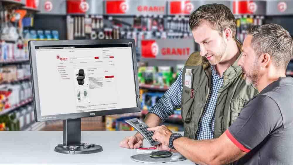To mænd står ved en disk og ser på en computer, der viser en hjemmeside med produkter.