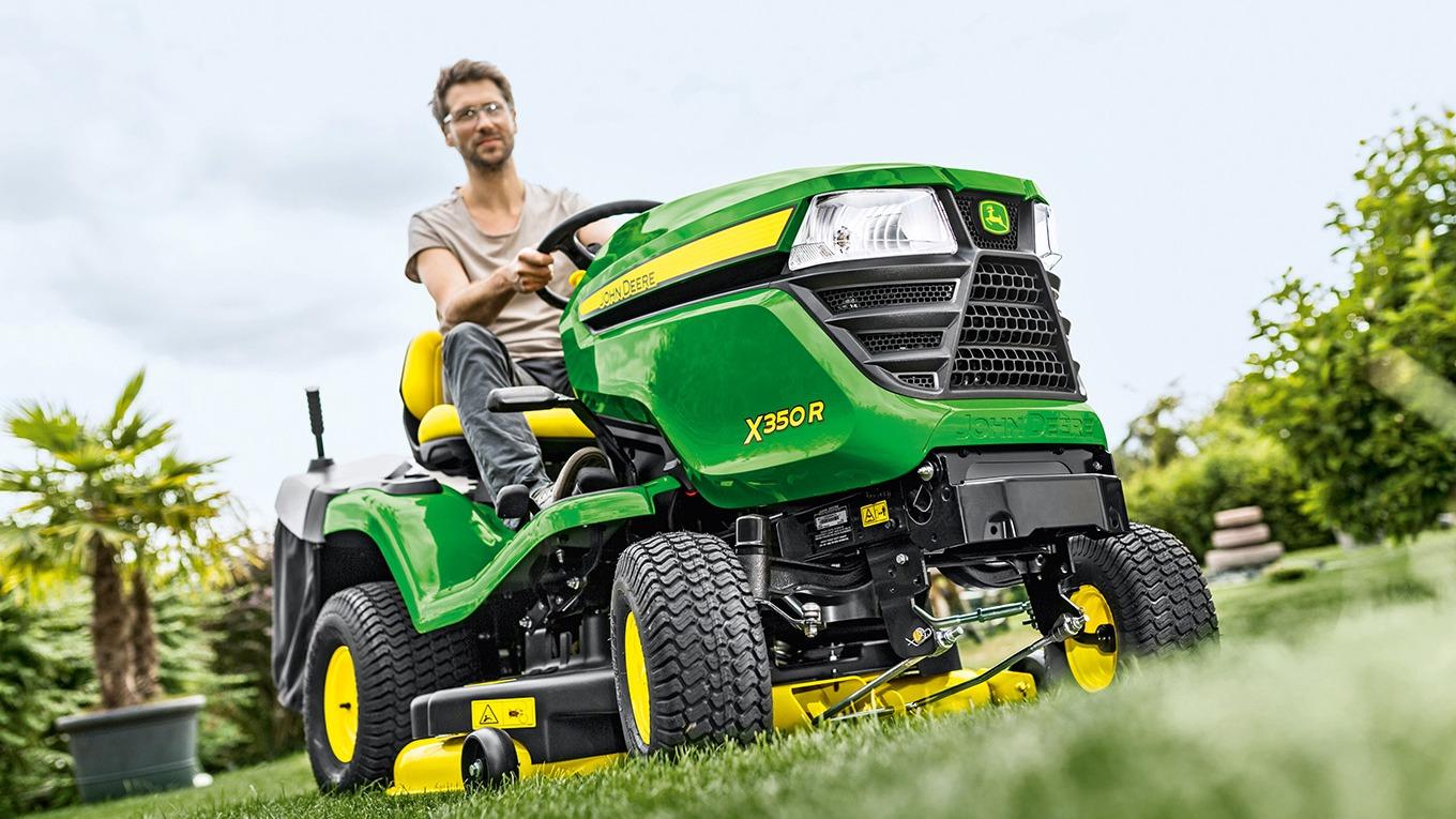 En mand kører en John Deere plæneklipper i en grøn have med træer og buske.