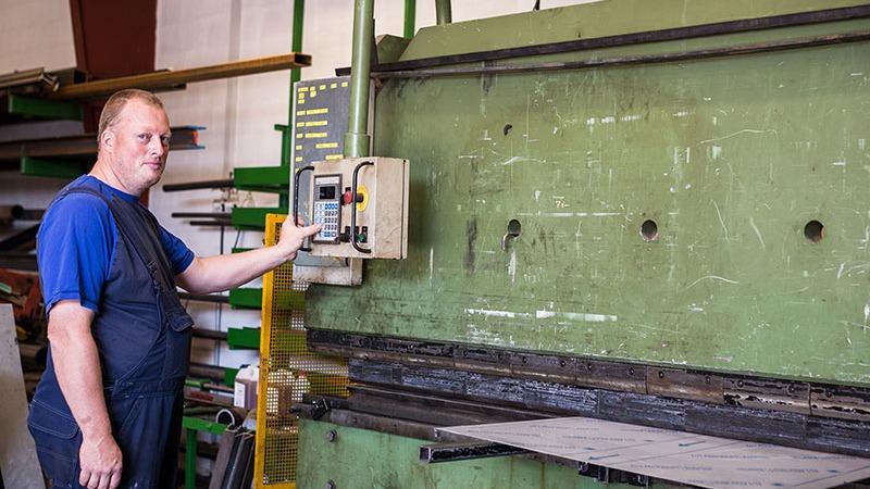 En mand betjener en stor metalpresser i et værksted, omgivet af metalplader og værktøj.