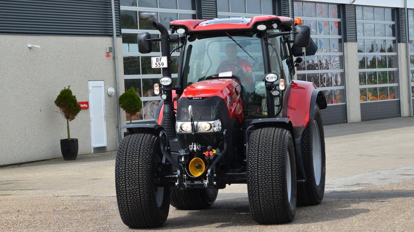 Rød traktor parkeret foran en bygning med store vinduer og planter i potter.