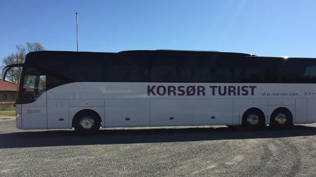 En turistbus fra Korsør med tydelig skrift på siden, parkeret på en grusvej under en klar blå himmel.