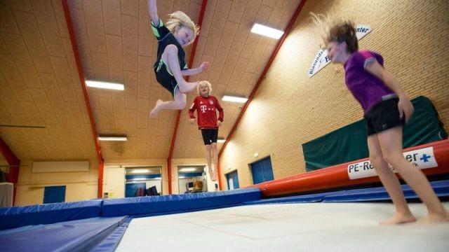Børn der hopper på en trampolin i en gymnastiksal, med glade ansigtsudtryk og energiske bevægelser.