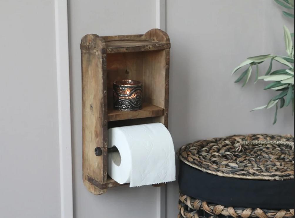 En væghængt toiletpapirholder i træ med en hylde til en dekorativ krukke og en kurv i baggrunden.