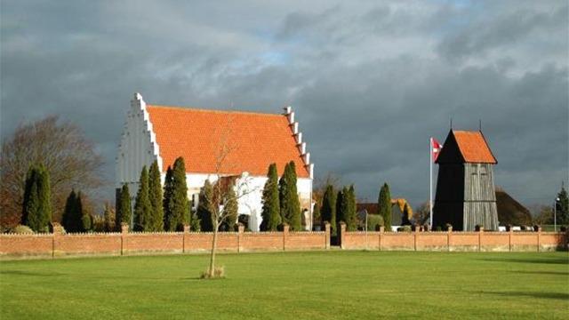 En hvid kirke med rød tag og en sort klokketårn omgivet af grønne træer og en græsplæne.