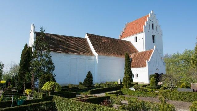 En hvid kirke med rød tagsten omgivet af velplejede haver og træer under en klar blå himmel.