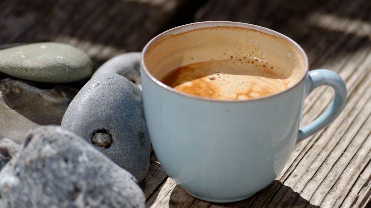 En blå kop med kaffe står på et træbord omgivet af grå sten.