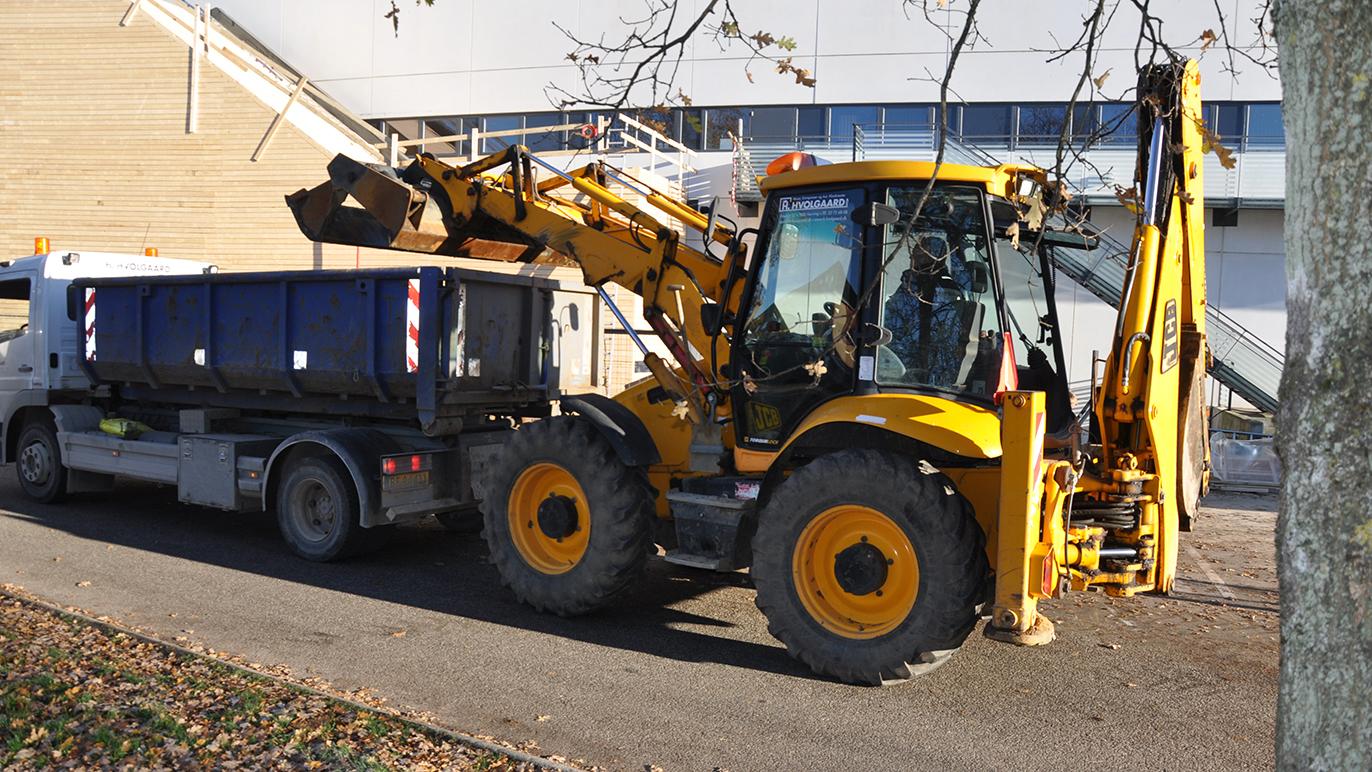 Gul gravemaskine parkeret ved en lastbil med en affaldscontainer i en byggeplads.