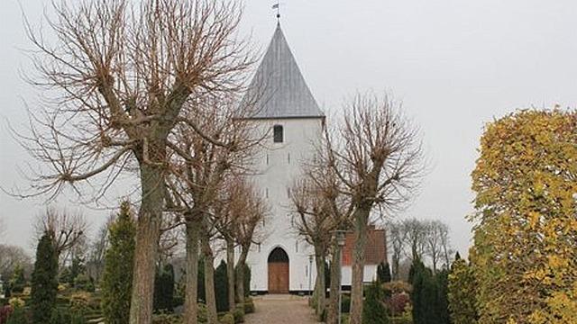 En hvid kirke med spidst tag omgivet af nøgne træer og en velholdt have i efterårsfarver.