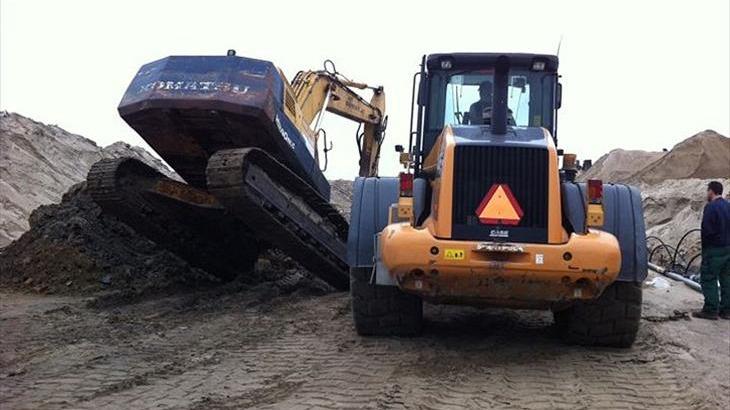 En gravemaskine og en gummihjulslæsser arbejder på en byggeplads med sand og grus.