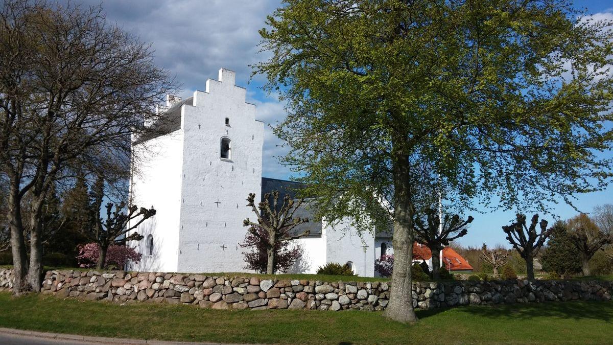 Hvidt hus med skråtag omgivet af træer og en stenmur under en blå himmel med skyer.