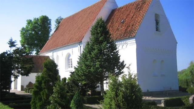 En hvid kirke med rødt tag omgivet af grønne træer og velplejede buske i solskin.