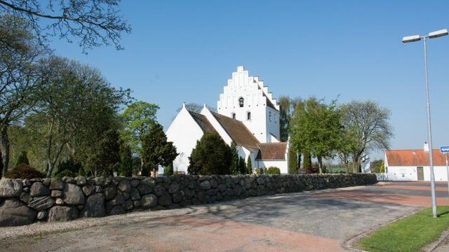 En hvid kirke med en spids tagform, omgivet af træer og en stenmur, beliggende ved en vej under en klar blå himmel.