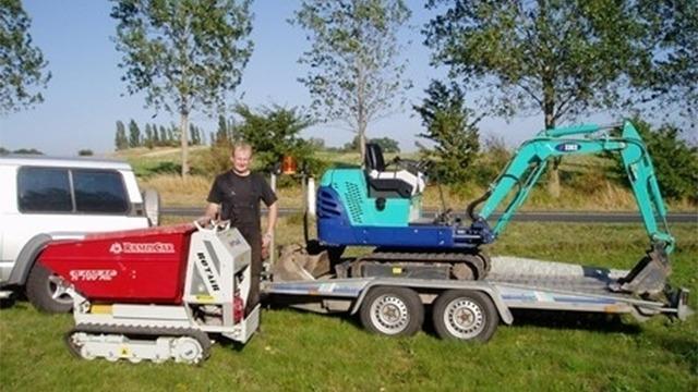 En mand står ved siden af en lille gravemaskine og en trailer med en større gravemaskine på en græsplæne.
